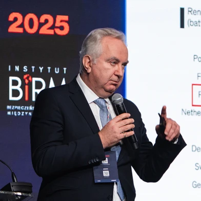 Osoba w formalnym ubraniu mówiąca do mikrofonu, z aparaturą audio w tle. Zdjęcie z konferencji lub wydarzenia w 2025 roku.