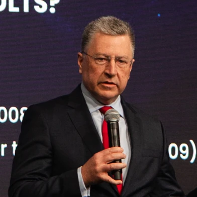 Mężczyzna w garniturze z okularami, przemawiający na wydarzeniu. Ma na sobie marynarkę i kołnierzyk na szyi.