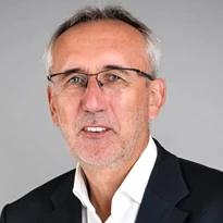 A man in formal wear with eyewear, touching his chin and cheek, with a raised eyebrow and a serious expression.