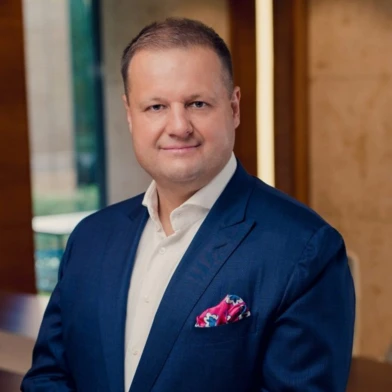 Чоловік у піджаку стоїть з посмішкою на обличчі, рукав піднятий до його чола. Стильний одяг, комір, видно плече.