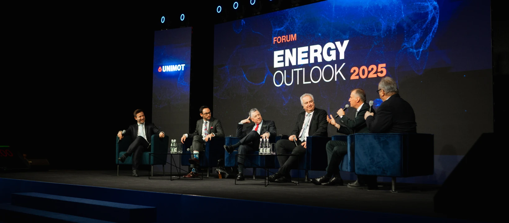 Pracownik biurowy w garniturze przemawia na scenie podczas konwencji technologicznej. Logo wydarzenia "UNIMOT" jest wyraźnie widoczne w tle.