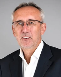 A man in formal wear with eyewear, touching his chin and cheek, with a raised eyebrow and a serious expression.
