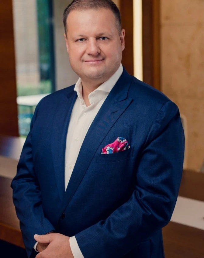 A man in a blazer standing with a smile on his face, his sleeve pushed up to his forehead. Formal wear, collar, shoulder in shot.