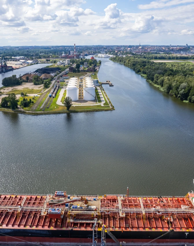 Statek transportowy przemierza wąską drogę wodną otoczoną drzewami i budynkami. Okręt transportowy prezentuje architekturę marynarską w działaniu.
