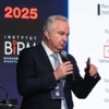 A person wearing formal wear speaking into a microphone, with audio equipment in the background. Image from a conference or event in 2025.
