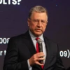 A man in formal wear with glasses, speaking at an event. He is wearing a suit and has a collar around his neck.
